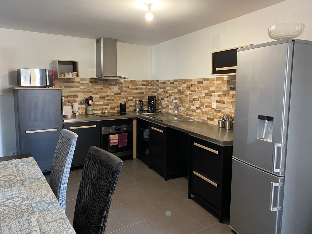 The kitchen features modern appliances, including a refrigerator and an oven. Stylish cabinetry is complemented by a tiled backsplash. A dining table with woven chairs is positioned to one side, creating an inviting space for meals and gatherings.