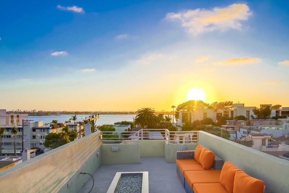A spacious rooftop deck features an orange sectional sofa arranged around a fire pit, with stunning views of a sunset over the water and surrounding landscape. The sky is filled with soft hues of blue and yellow, creating a serene atmosphere.