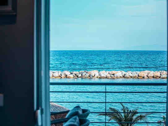 A clear view of the calm sea is framed by a doorway, showcasing the tranquil blue waters meeting a rocky breakwater. The horizon stretches into the distance, creating a peaceful coastal atmosphere.
