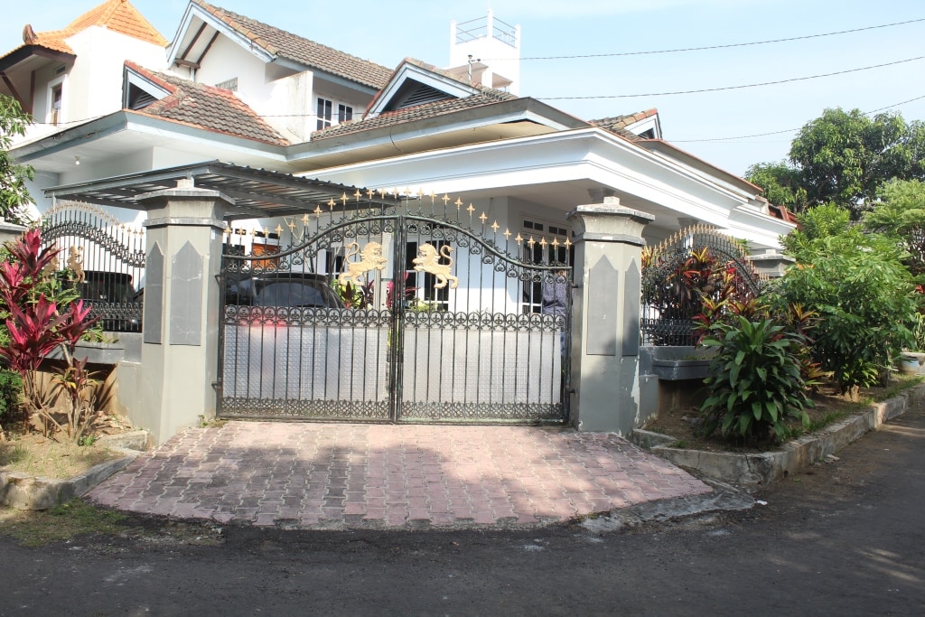 A gated entrance is featured, framed by a decorative wrought iron fence. The pathway is paved with bricks, leading to a spacious front yard adorned with lush greenery and vibrant plants. The structure behind the gate suggests a welcoming residential setting.