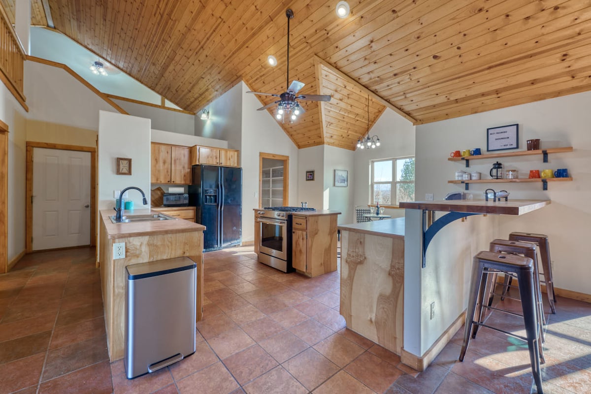 A warm kitchen environment features wood accents and high vaulted ceilings. Equipped with stainless steel appliances, including a refrigerator and gas range, the space offers a large island with bar seating and ample natural light from the windows.