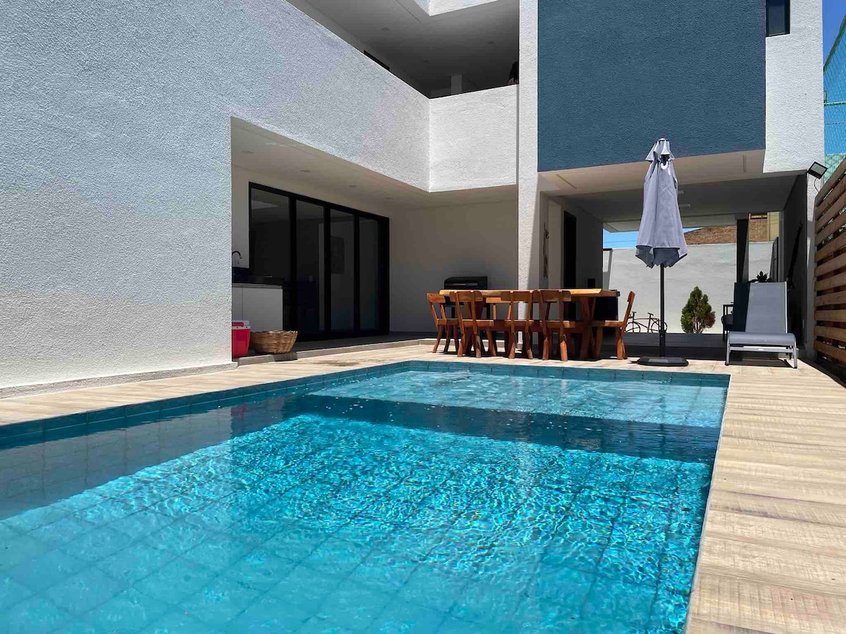 An outdoor area features a bright blue pool surrounded by a wooden deck. A spacious dining table with multiple chairs sits nearby, and a sun umbrella provides shade. The modern architecture showcases large glass doors leading to the interior spaces.