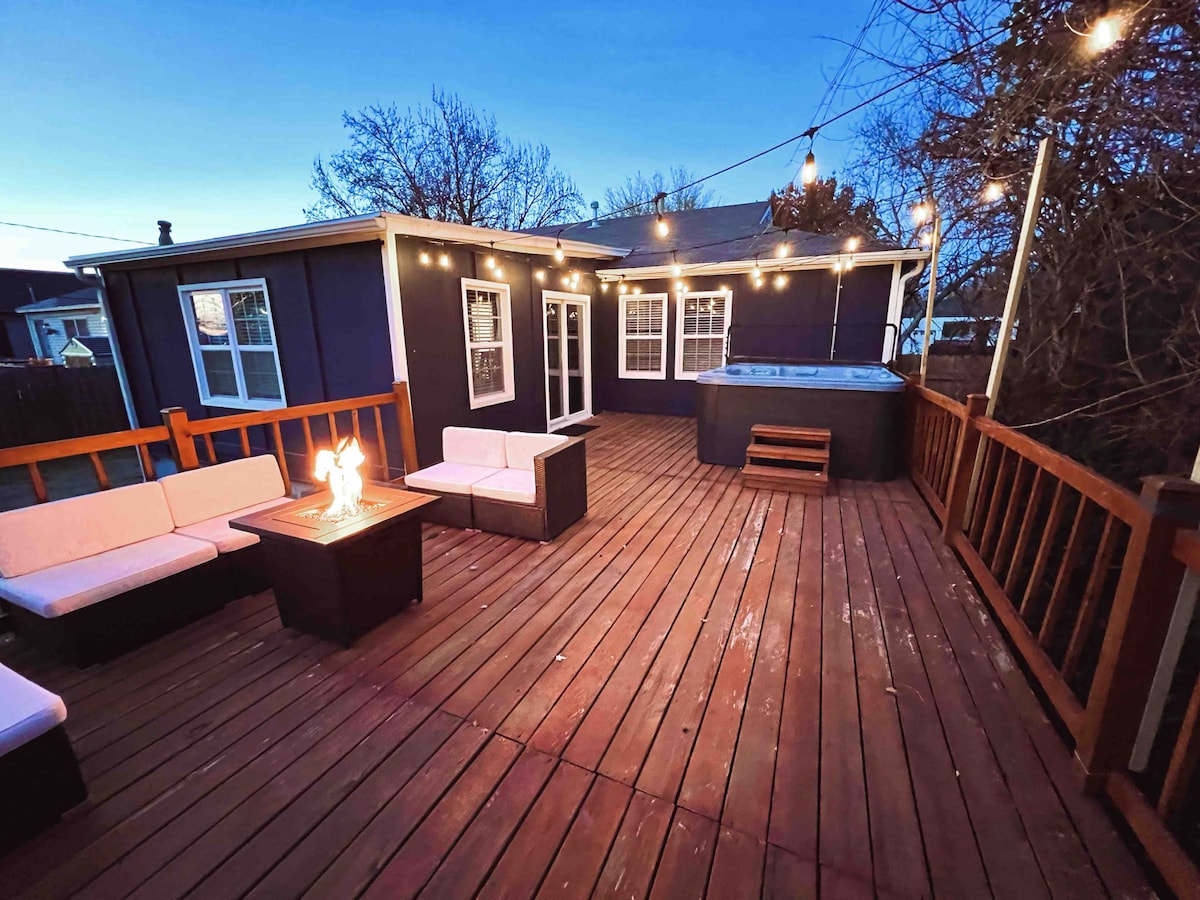 A spacious wooden deck is illuminated by string lights, creating an inviting atmosphere. Comfortable seating is set around a fire pit, and a hot tub is visible in the corner, offering a relaxing outdoor space for gatherings.