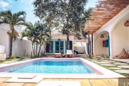 A serene outdoor space features a private pool surrounded by lush greenery. Comfortable lounge chairs are positioned beside the water, with colorful doors and windows visible on the adjacent building. Natural wood elements enhance the relaxed atmosphere.