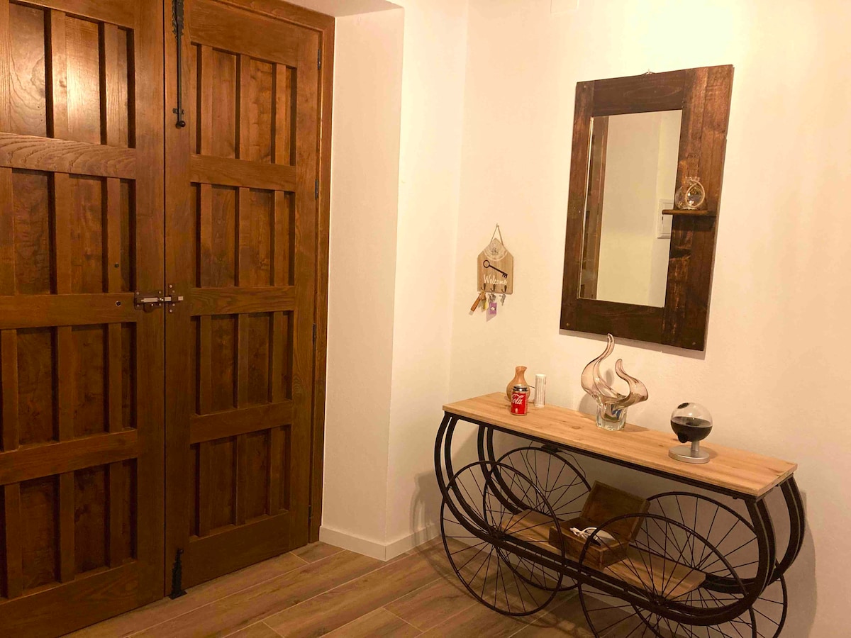 An inviting entryway features wooden double doors and a console table with a minimalist design. A round mirror reflects light, while decorative items and a small vase add subtle elegance to the space. Warm lighting enhances the cozy atmosphere.