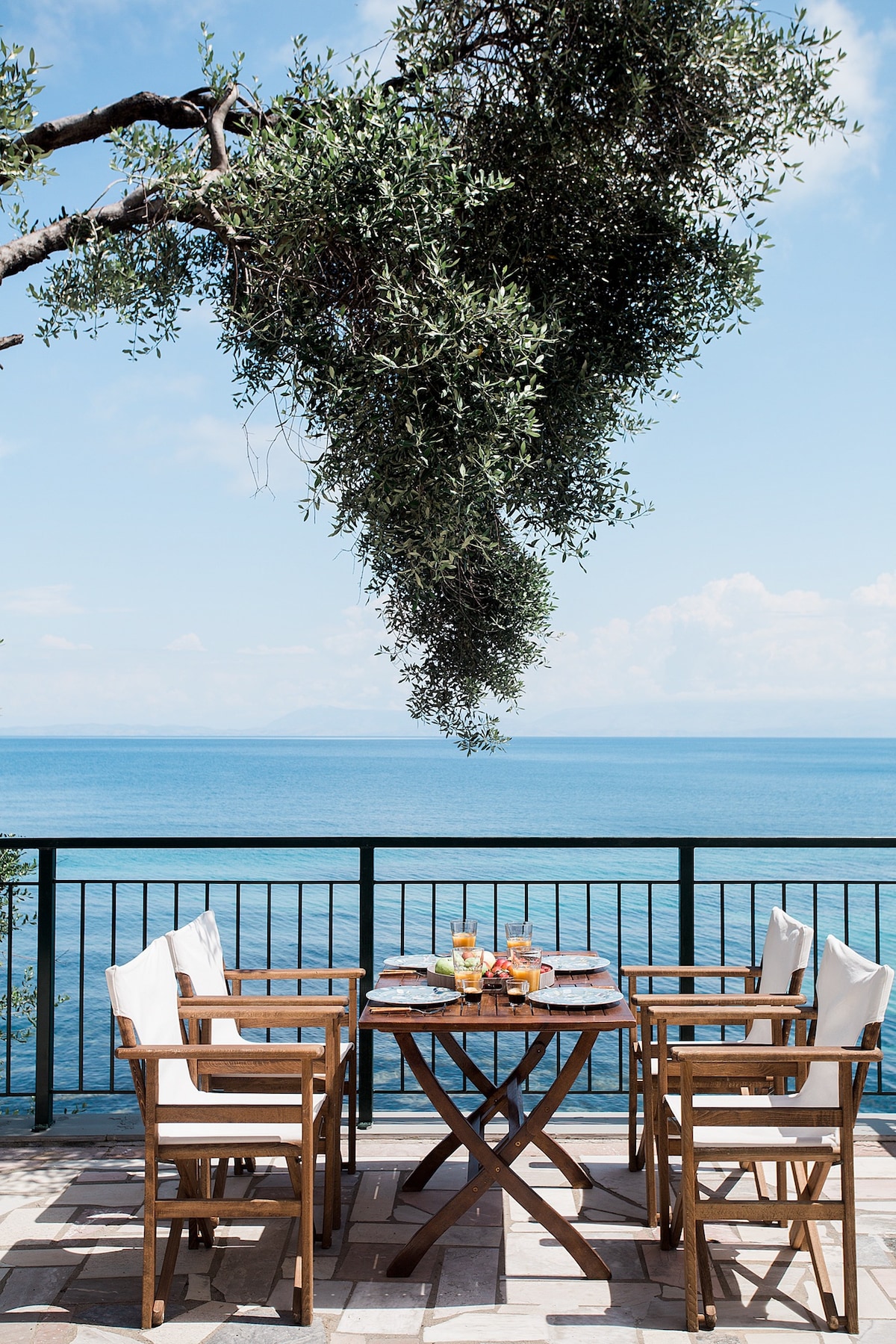 An outdoor dining area features a wooden table surrounded by four chairs, set against a backdrop of the Ionian Sea. The table is adorned with breakfast items and drinks. A leafy olive tree hangs above, providing a touch of natural shade.