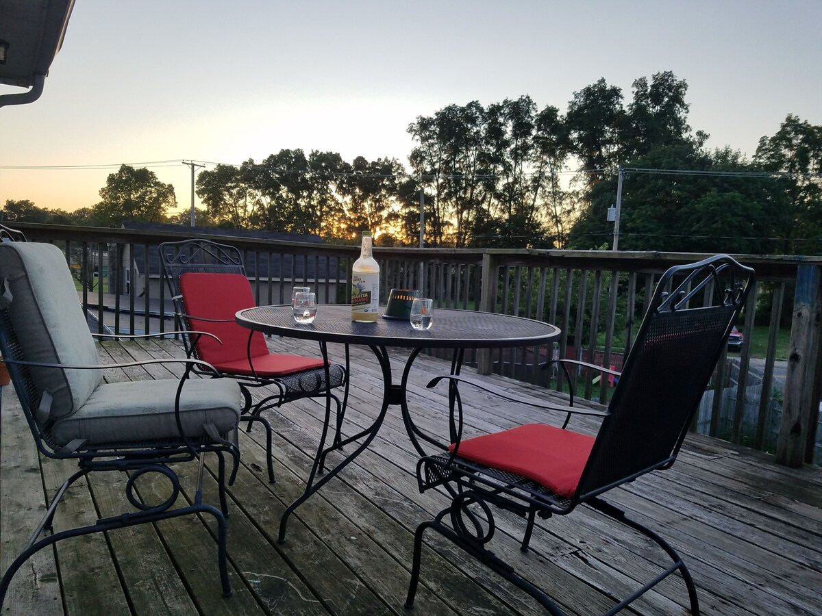 An outdoor deck area features a circular table with four chairs, two of which have red cushions. A bottle sits on the table alongside empty glasses. The backdrop showcases trees against a sunset sky, creating a serene setting for relaxation.