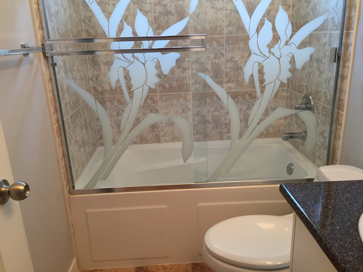 A clean bathroom features a glass shower enclosure with floral designs. A bathtub is visible alongside a toilet, with a dark countertop presenting a modern aesthetic. Neutral wall tones complement the tiled flooring and decorative elements.