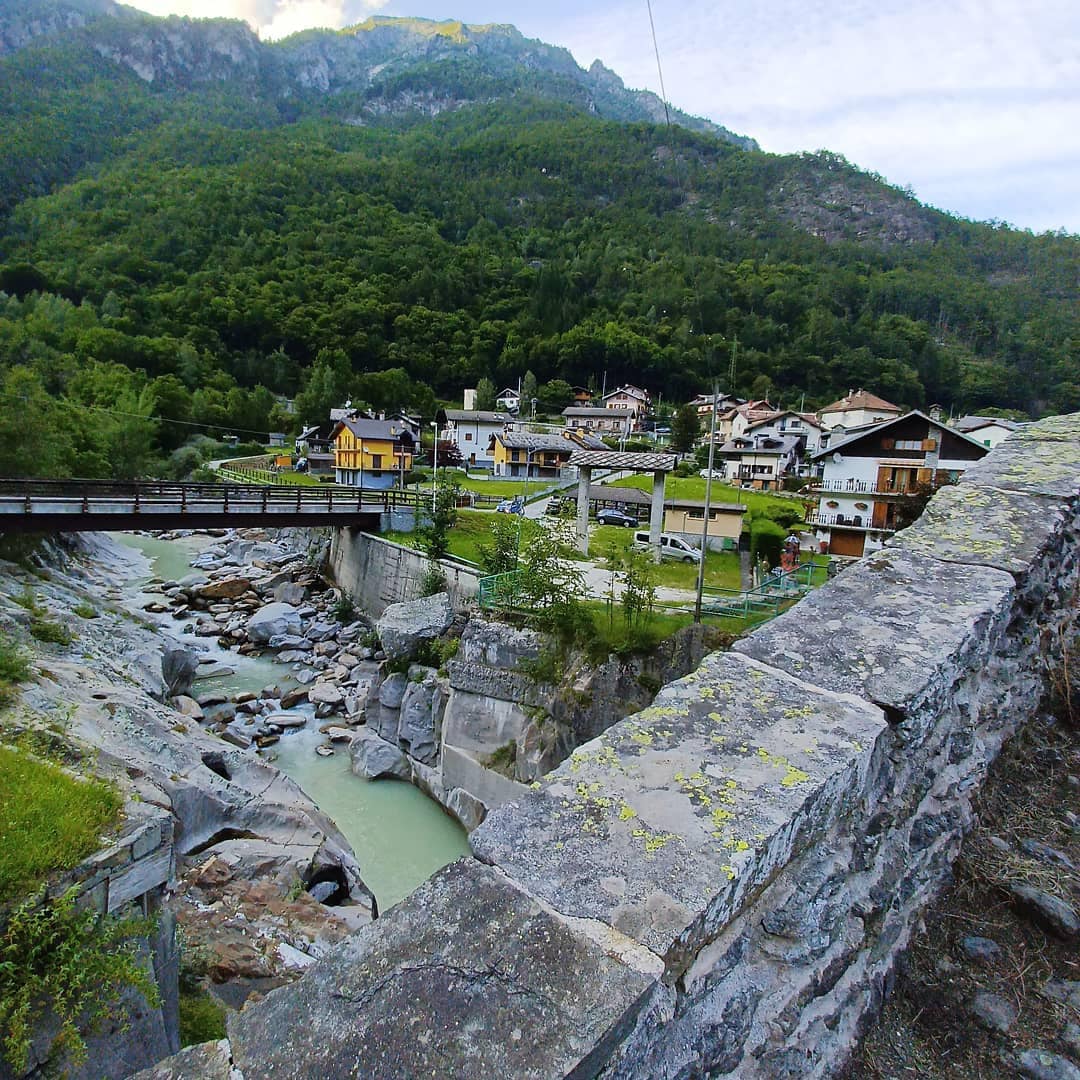 A view of a picturesque mountain village nestled in a lush valley, with buildings featuring varied architectural styles. A stone bridge spans a river, while the surrounding greenery and rocky terrain highlight the natural beauty of the area. Mountains loom in the background.