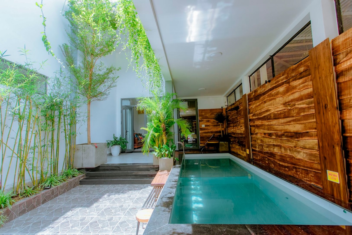 A serene swimming pool is framed by natural wooden panels, creating a tranquil atmosphere. Lush greenery, including tall plants and bamboo, complements the space. The pool reflects the surrounding light, enhancing the calm setting suitable for relaxation.