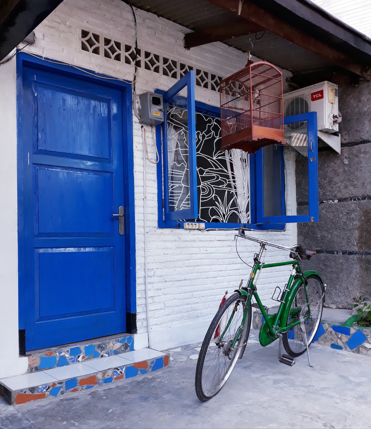 A vibrant blue door is set against a textured white wall, framed by a cage hanging from an open window. A green bicycle is parked on the concrete surface, enhancing the inviting entrance of the pavilion.