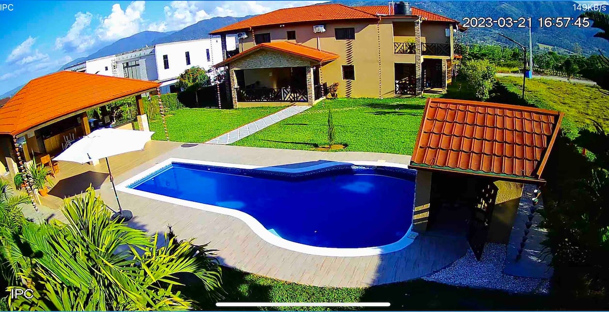 The outdoor area features a large swimming pool with a clear blue surface, surrounded by a spacious deck. Several green lawns and landscaped areas are visible, along with a gazebo and an outdoor dining space under an umbrella. The background showcases the serene environment and nearby structures.
