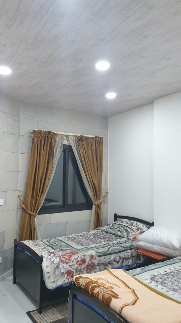 The sleeping area features two single beds adorned with patterned blankets. A decorative window with dark curtains allows natural light to filter in. Warm-toned curtains frame the window, complementing the neutral wall color and modern ceiling lights.