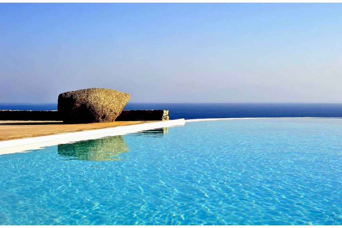 An infinity swimming pool reflects the clear sky above, with gentle waves visible in the sea beyond. A rounded stone structure is positioned at the pool's edge, adding a natural element. The tranquil blue water seamlessly blends into the horizon.