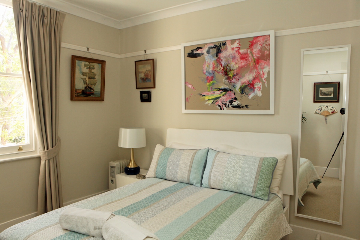 The softly hued bedroom features a spacious bed with a striped coverlet and two pillows. A framed artwork hangs above the bed, while a full-length mirror reflects the room's light. Light curtains frame the window, and a bedside lamp provides additional illumination.