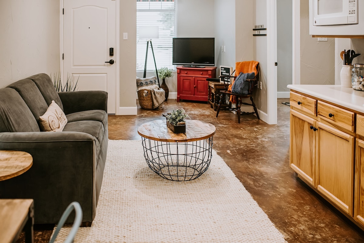 A cozy living area is presented with a dark gray sofa and a circular wire coffee table. Light streams in through a window adorned with simple treatments. A red cabinet contrasts with the beige walls, and a rustic rocking chair adds charm. The floor features a textured finish.