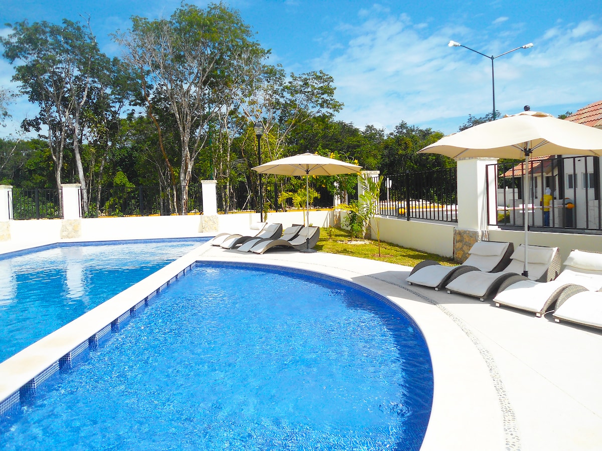 A clear blue pool is highlighted under bright skies, surrounded by green trees. Comfortable lounge chairs are arranged along the poolside, with large umbrellas providing shade. The serene environment invites relaxation in a spacious outdoor area.