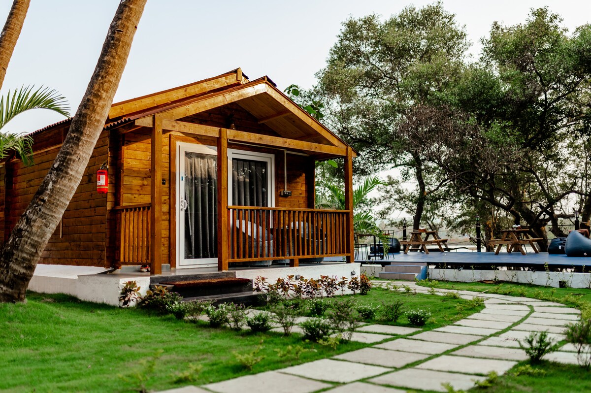 A wooden chalet is positioned adjacent to a lush garden, featuring a porch with railings. The pathway is lined with stepping stones, leading to an outdoor sitting area surrounded by trees. The setting offers a sense of privacy and natural beauty.