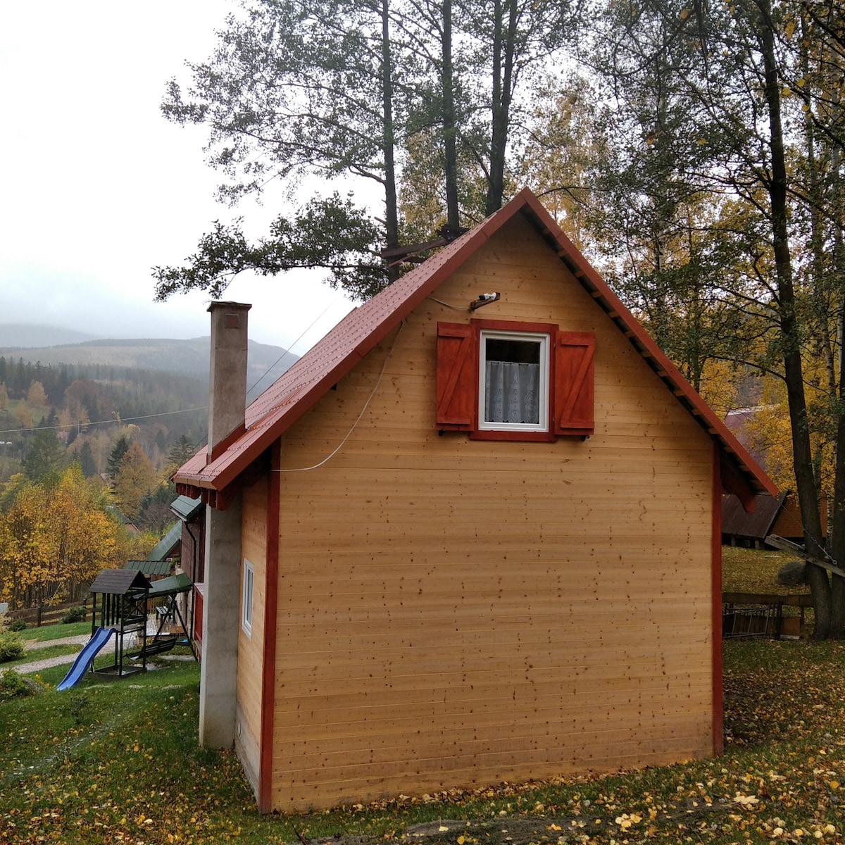 The exterior of a light wooden cottage is visible, featuring a sloped red roof. A small window with white curtains is positioned on the gabled side. Surrounding trees showcase autumn foliage, while a grassy area includes a slide and wooden seating in the background.