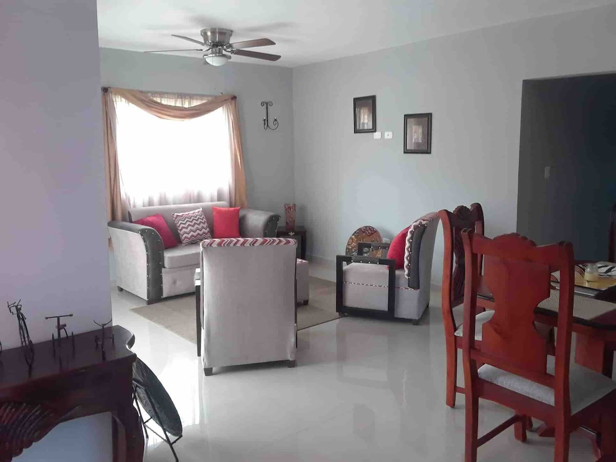 The living area features a combination of light-colored sofas with accent cushions and a matching coffee table. Natural light is welcomed through large windows, complementing the ceramic flooring. A dining table with wooden chairs is positioned nearby, enhancing the space's functionality.