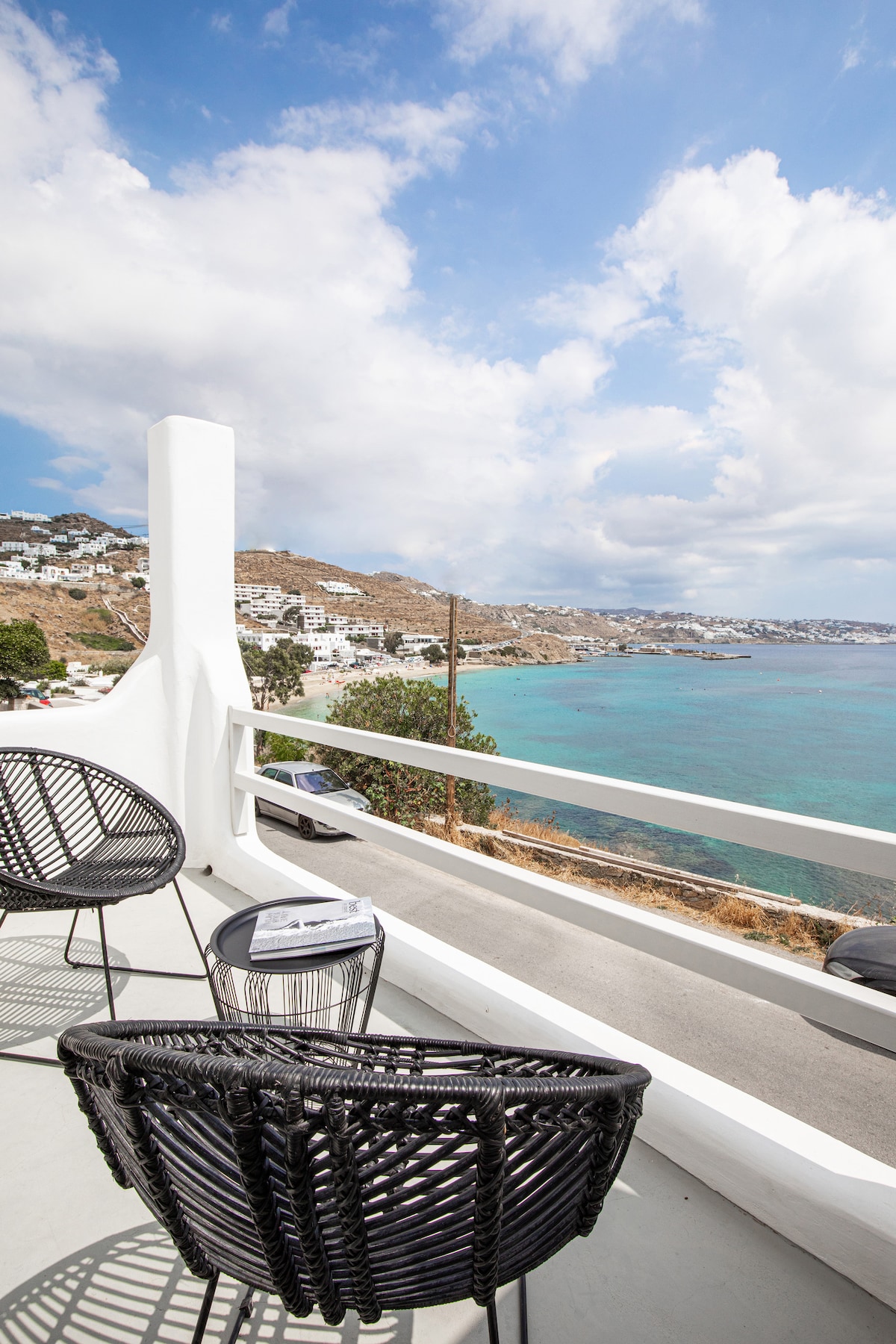 A balcony showcases a pair of modern black woven chairs positioned for optimal views of the Aegean Sea. The turquoise waters stretch across the horizon, framed by gentle coastal hills under a partly cloudy sky.