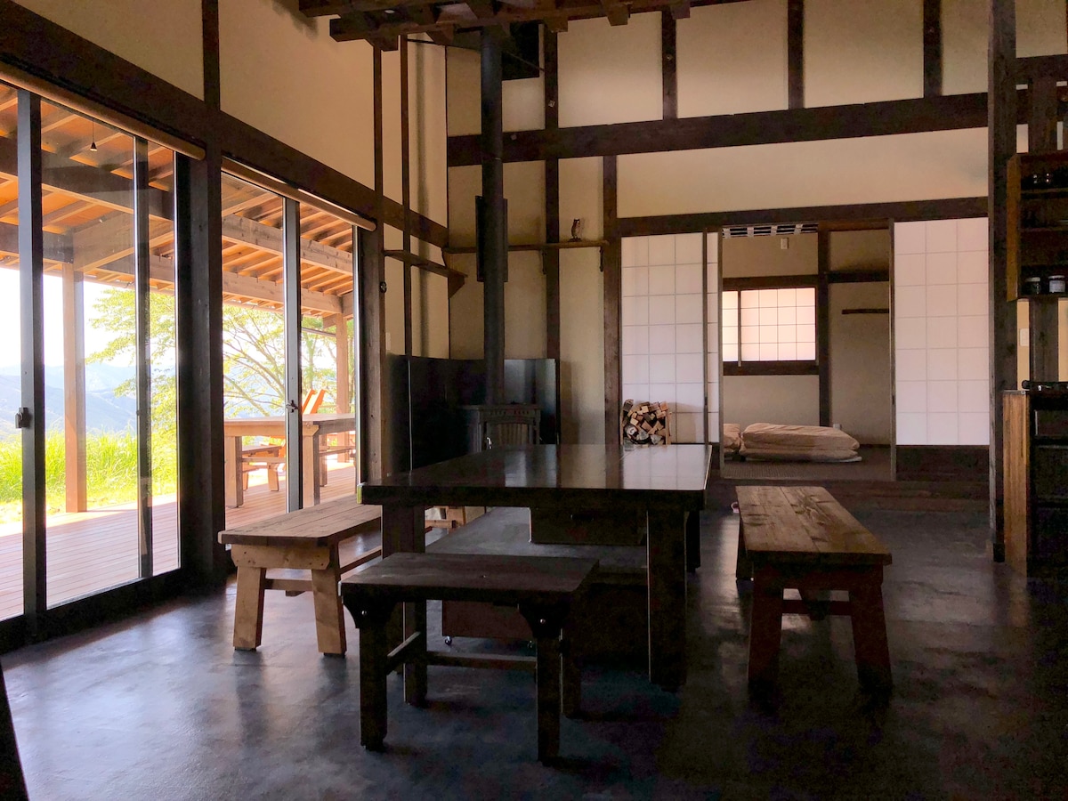 An open interior features wooden beams and expansive glass doors that provide a view of the surrounding landscape. A central wooden table is surrounded by benches, and a traditional Japanese-style room is visible in the background, enhancing the rustic charm of the space.