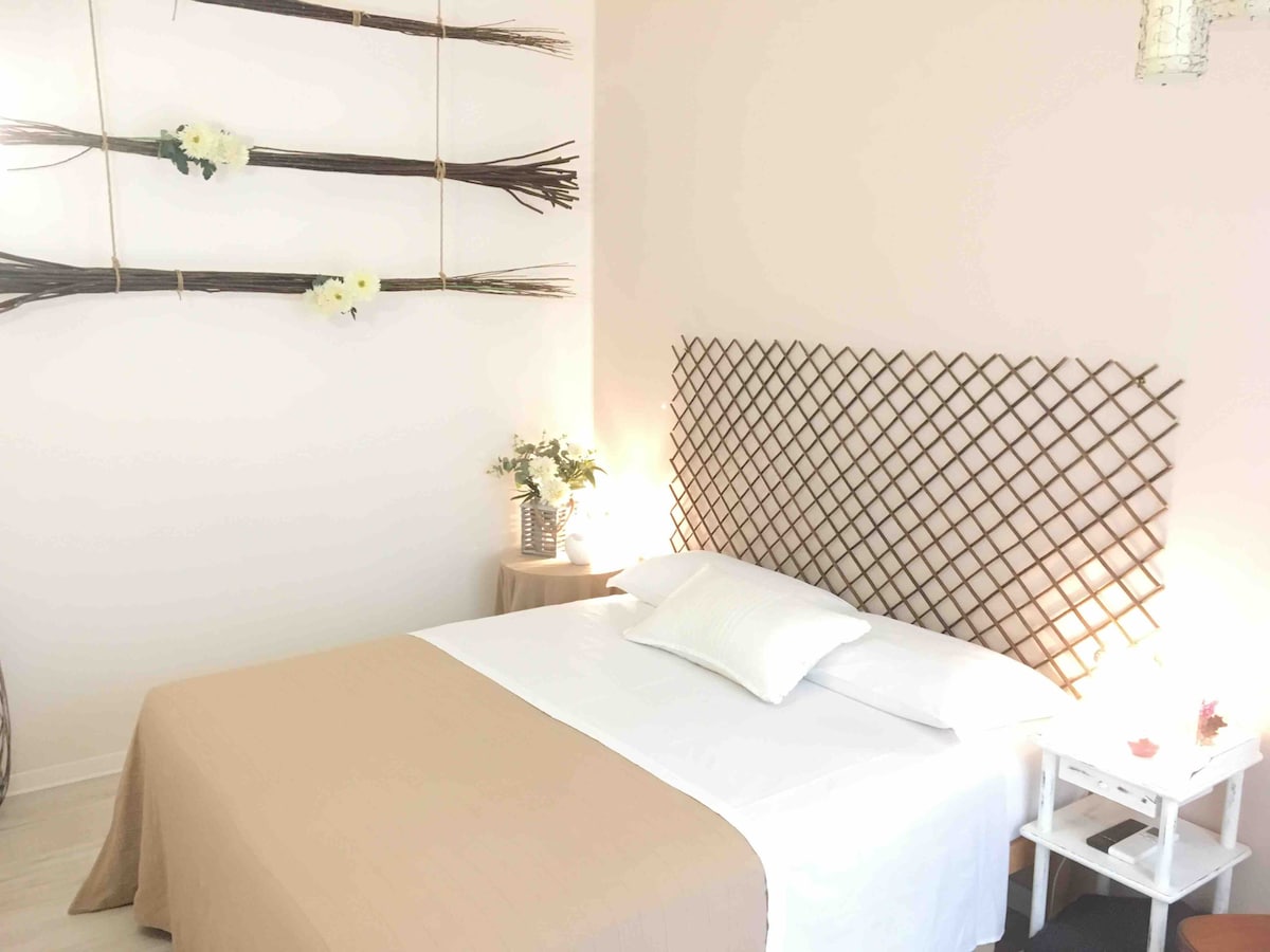 A softly lit room features a spacious bed with a neutral-toned blanket. Decorative wall elements create a simple, stylish backdrop. A bedside table holds a small vase of flowers, while a light fixture adds a warm glow to the ambiance.