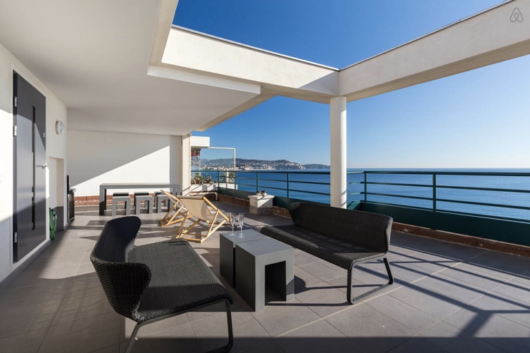 An expansive terrace features modern outdoor furniture, including a seating area with two chairs and a table. Breathtaking sea views are visible beyond a glass railing, complemented by clear blue skies. The space is designed for relaxation and outdoor enjoyment.