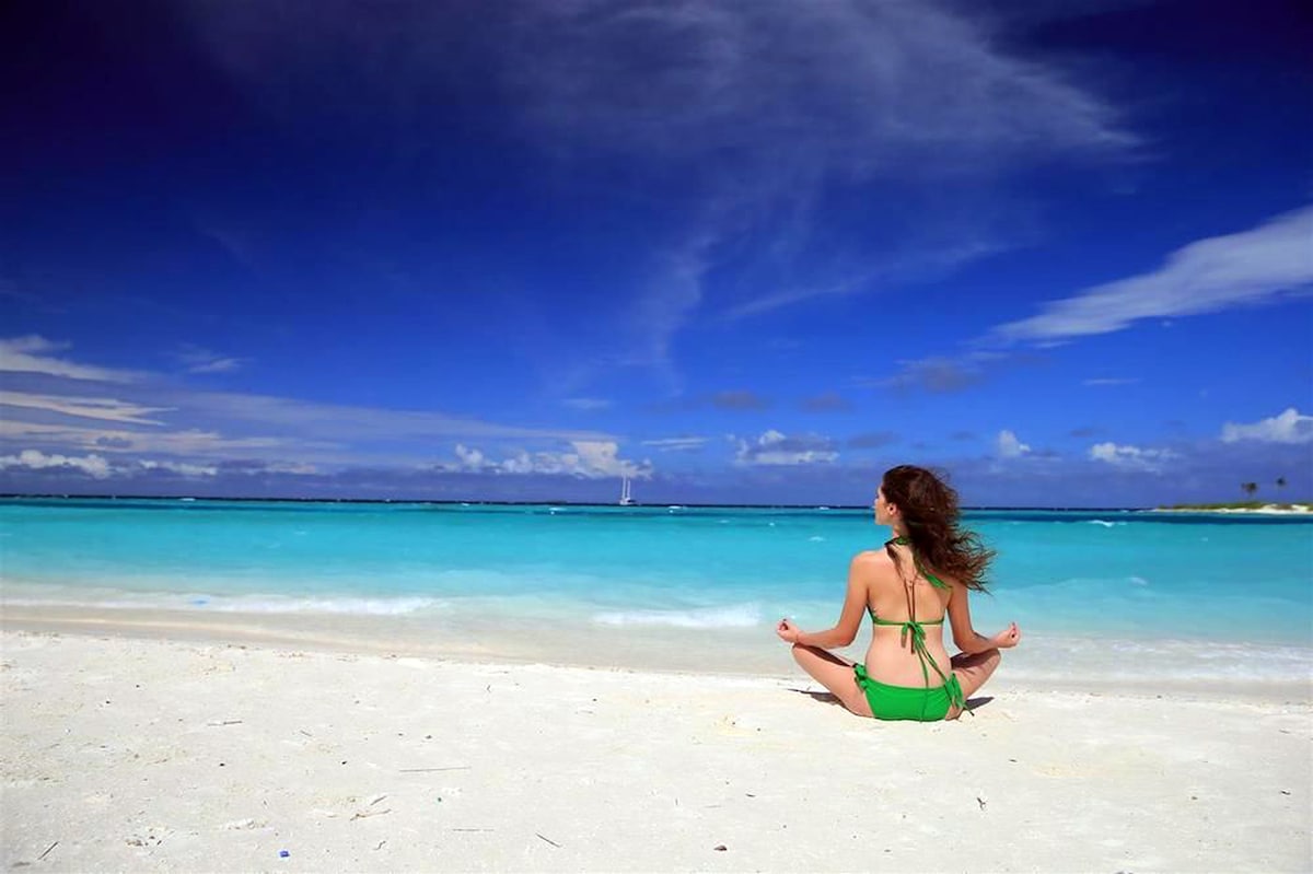 A woman is seated on soft, white sand, facing the serene turquoise water. The expansive sky above is bright blue, with wispy clouds. Gentle waves touch the shore, and a sailboat can be seen in the distance, contributing to a tranquil beach atmosphere.
