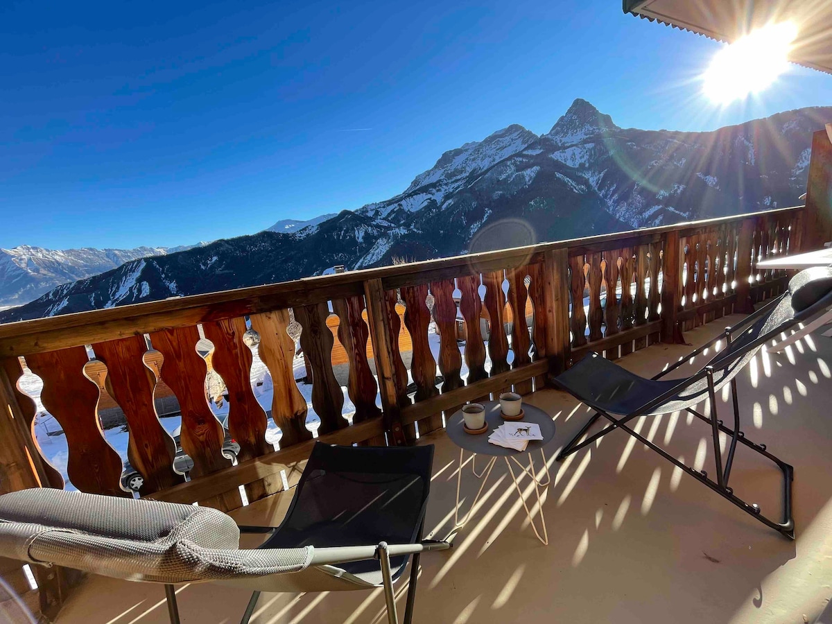 A balcony is presented with views of surrounding mountains and a clear blue sky. Two lounge chairs and a small table are positioned in the sunlight. The wooden railing adds a rustic touch to the outdoor space.