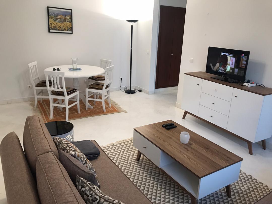 A modern living area is presented, featuring a comfortable sofa and a wooden coffee table. A television is positioned on a stylish white cabinet. A white dining table surrounded by four chairs is highlighted, with a piece of art adorns the wall above the table.