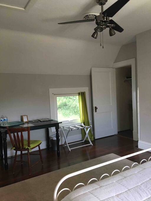 Large desk, luggage rack, ceiling fan and closet with hangers.