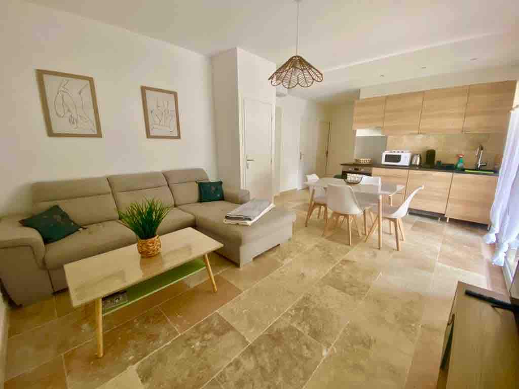 A light-filled living area showcases a spacious gray sofa accompanied by decorative cushions. A wooden coffee table sits in front, while a dining set with four chairs is nearby. The kitchen area features modern cabinetry and appliances. Neutral tones highlight the inviting atmosphere.