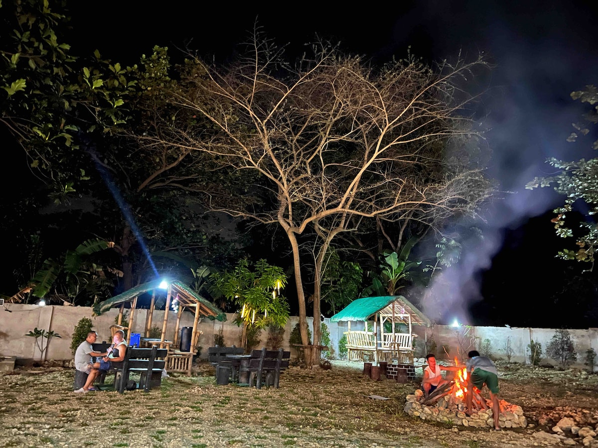 A spacious outdoor area is illuminated by soft lighting, featuring a large, bare tree amidst greenery. Campfires are tended by guests while seating arrangements with tables and chairs are visible in the background. Two cozy gazebos provide additional gathering spots.