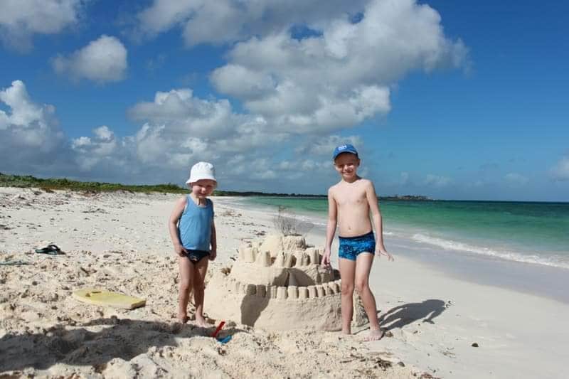 Two children stand on a sandy beach beside a large sandcastle. The beach features soft, light-colored sand and gentle waves in the background. A clear blue sky with fluffy white clouds stretches overhead, enhancing the serene coastal atmosphere.