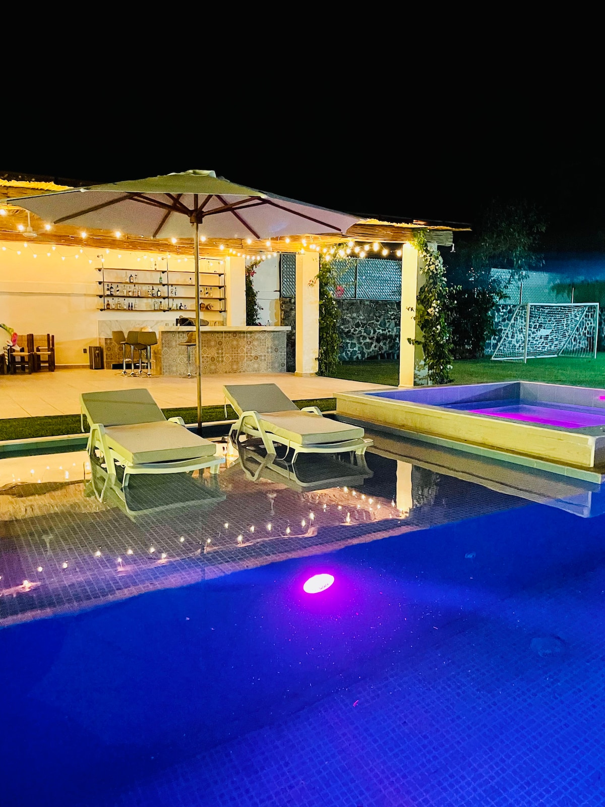 The illuminated pool area features comfortable lounge chairs beside the water. A cozy jacuzzi sits adjacent, glowing with soft purple lighting. Twinkling string lights enhance the outdoor space, complemented by a shaded area with a table and chairs in the background.