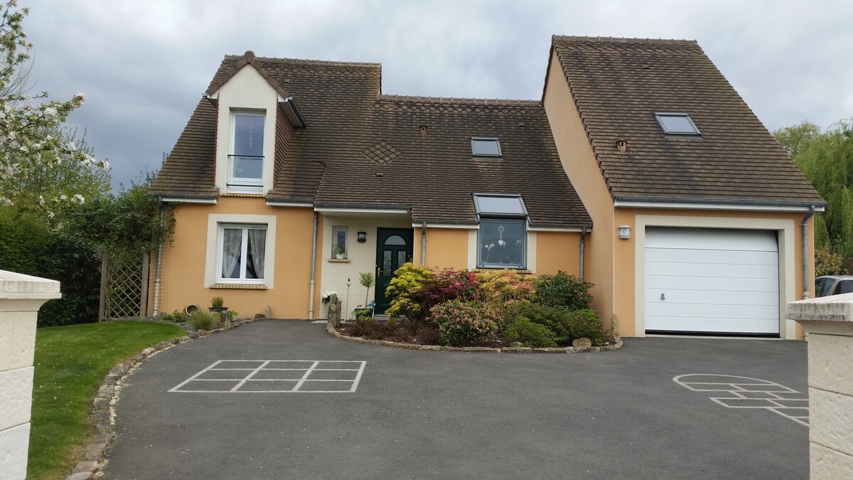 A spacious two-story house features a beige exterior with a sloped roof. A driveway is visible, bordered by well-maintained gardens with colorful plants. A large garage door is present on the right side, and several windows allow natural light to enter the home.