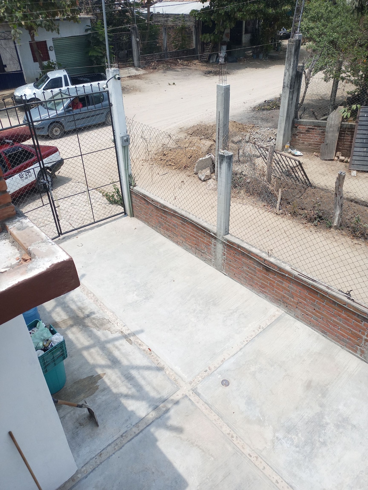 An exterior view showcases a concrete pathway leading to a fenced area with a gate. The surrounding environment captures a glimpse of the street with vehicles and an unfinished area visible in the background. The space appears tidy and well-maintained.