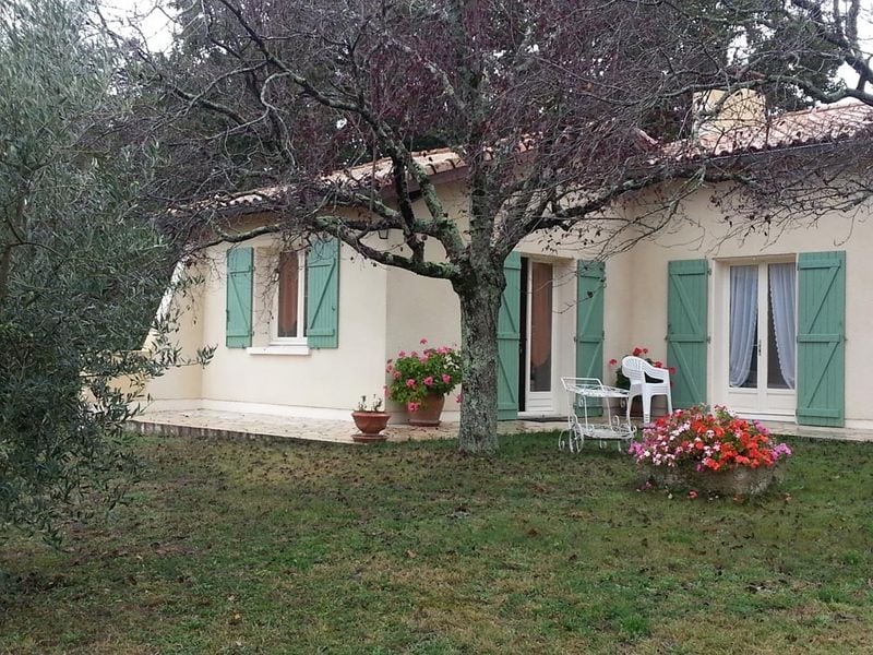 A single-story house features light-colored walls and green shutters, set against a lush garden. A flowering tree provides shade, while potted plants with vibrant blooms are positioned near the entrance. A small outdoor seating area is visible, enhancing the tranquil ambiance.