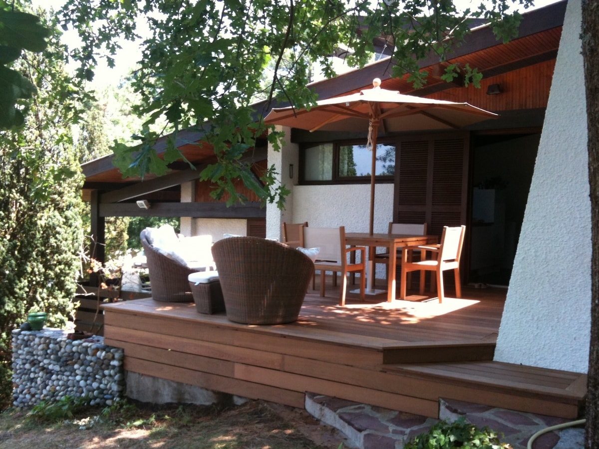 A wooden deck is featured, furnished with a dining table and chairs, along with two comfortable seating options. An umbrella provides shade, while greenery surrounds the area, creating a pleasant outdoor space. The structure presents a combination of textured walls and a sloped roof.