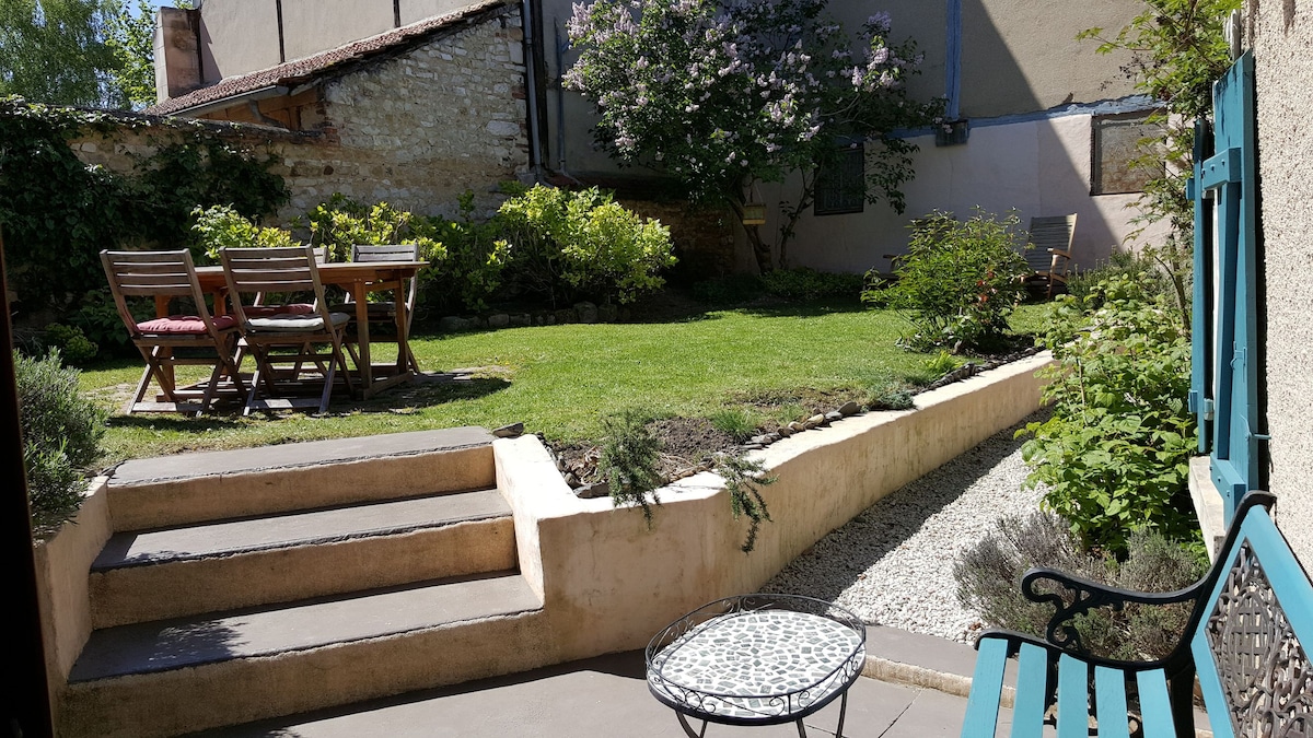 The garden is surrounded by greenery and features a well-maintained lawn. A wooden dining table with matching chairs provides an outdoor dining area. Decorative plants accentuate the space, and a small patio is accessible via steps leading down from the house.