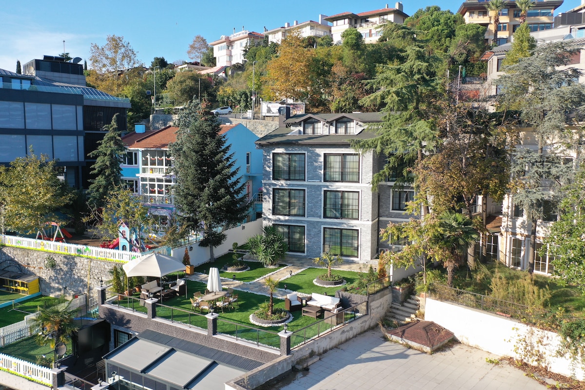 A spacious outdoor area showcases a mix of green lawns and landscaping surrounding a modern multi-story building. Seating areas and shaded umbrellas provide inviting spots for relaxation, while the backdrop of trees and nearby structures creates a tranquil environment.