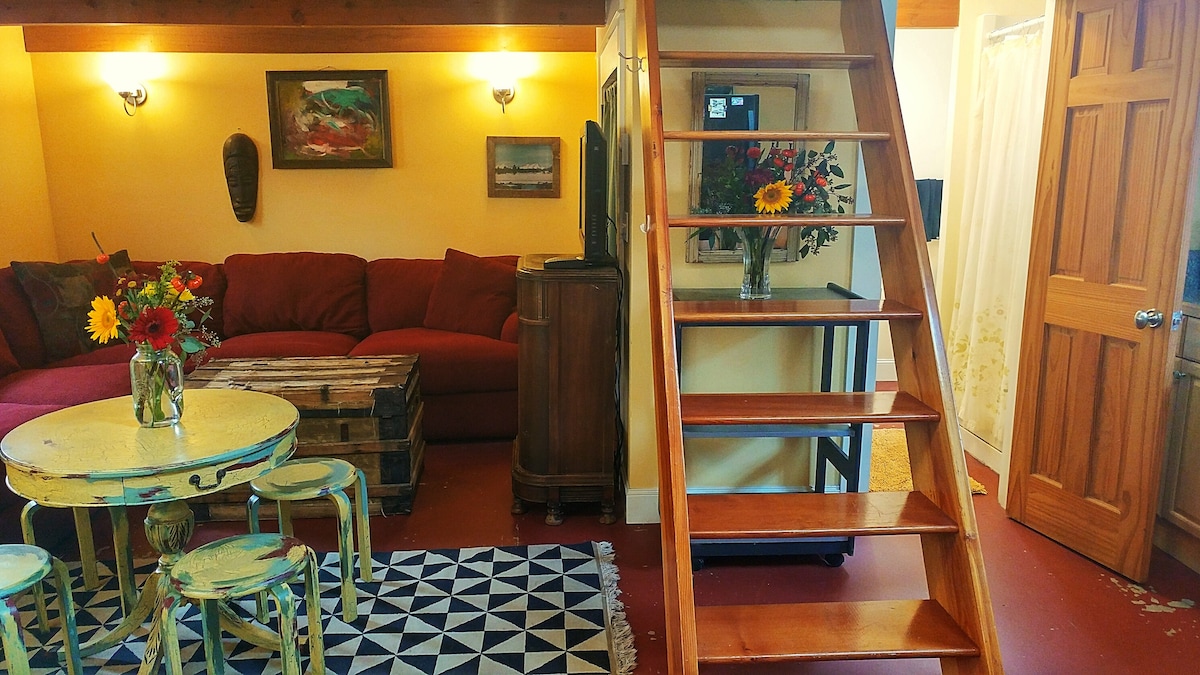 A cozy living area features a large red sectional sofa and a rustic wood coffee table adorned with colorful flowers. A patterned rug covers the floor, while a staircase leads to an upper loft space. Soft lighting enhances the inviting atmosphere.