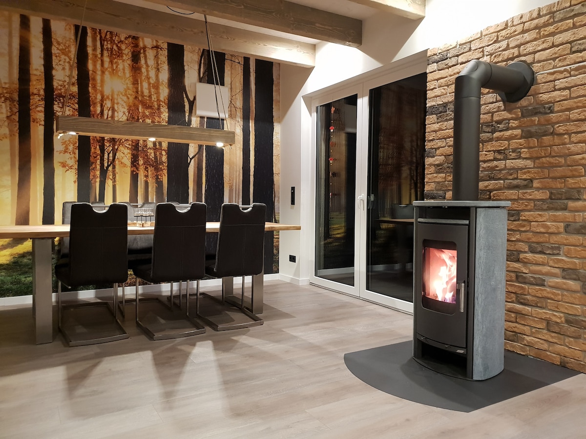 A dining area is highlighted by a large wooden table surrounded by six dark dining chairs. A modern wood stove on a circular mat emits gentle warmth beside a textured stone wall. Large glass doors provide access to the outside, with a forest mural enhancing the ambiance.
