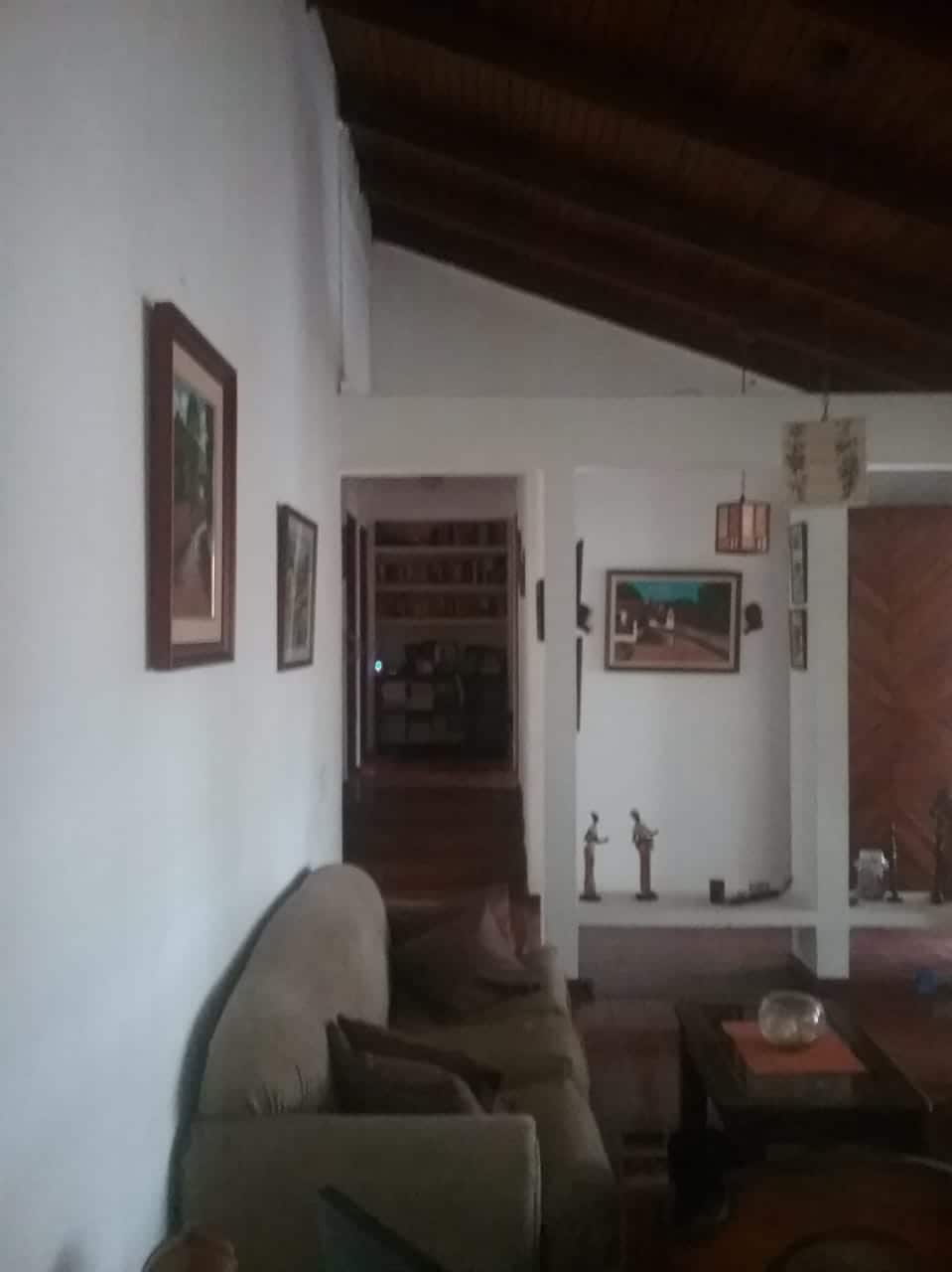 A spacious living area is featured, showcasing a neutral-colored sofa adorned with colorful cushions. Wall-mounted artwork decorates the white walls, and a wooden coffee table is positioned in front. Light enters from an adjacent hallway, where a library can be glimpsed.