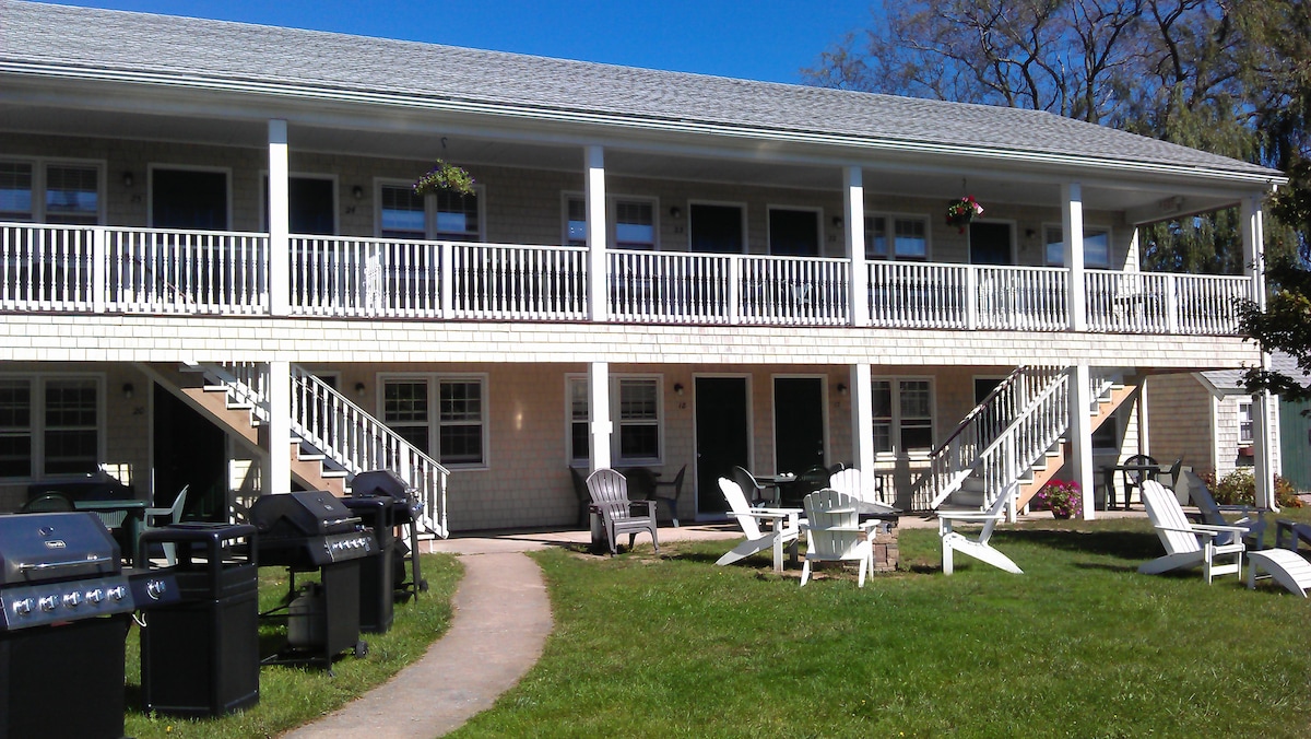 The exterior of a two-story building is displayed, featuring a row of doors and a covered balcony. Grills are available for use on a well-kept lawn, which is complemented by several adirondack chairs arranged for relaxation. The surrounding greenery enhances the outdoor space.
