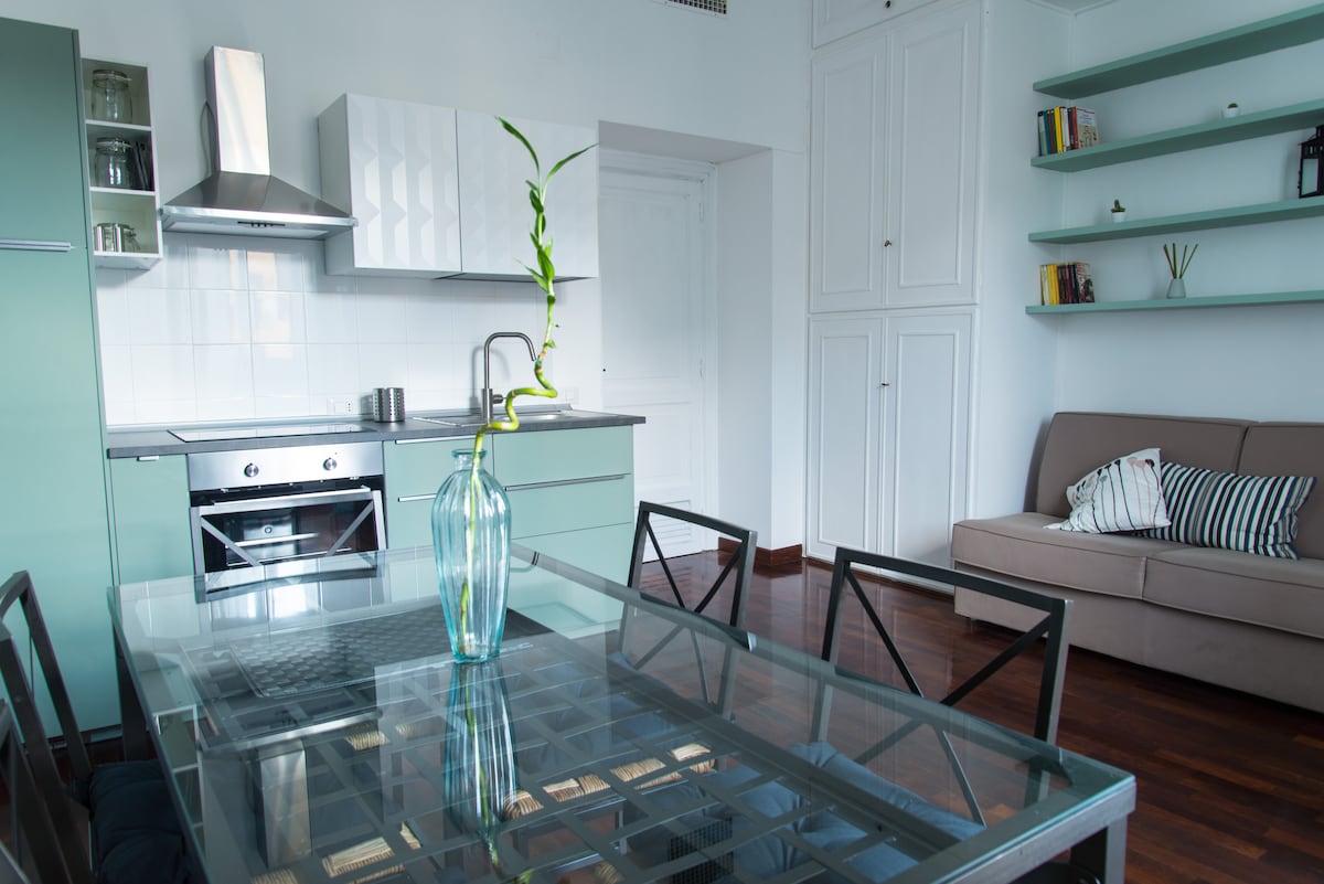 The kitchen area is equipped with modern appliances, featuring a stove, oven, and refrigerator. A dining table with black metal chairs is positioned nearby. A vase with greenery is centered on the table. A comfortable sofa is visible against the wall, with shelves displaying books.