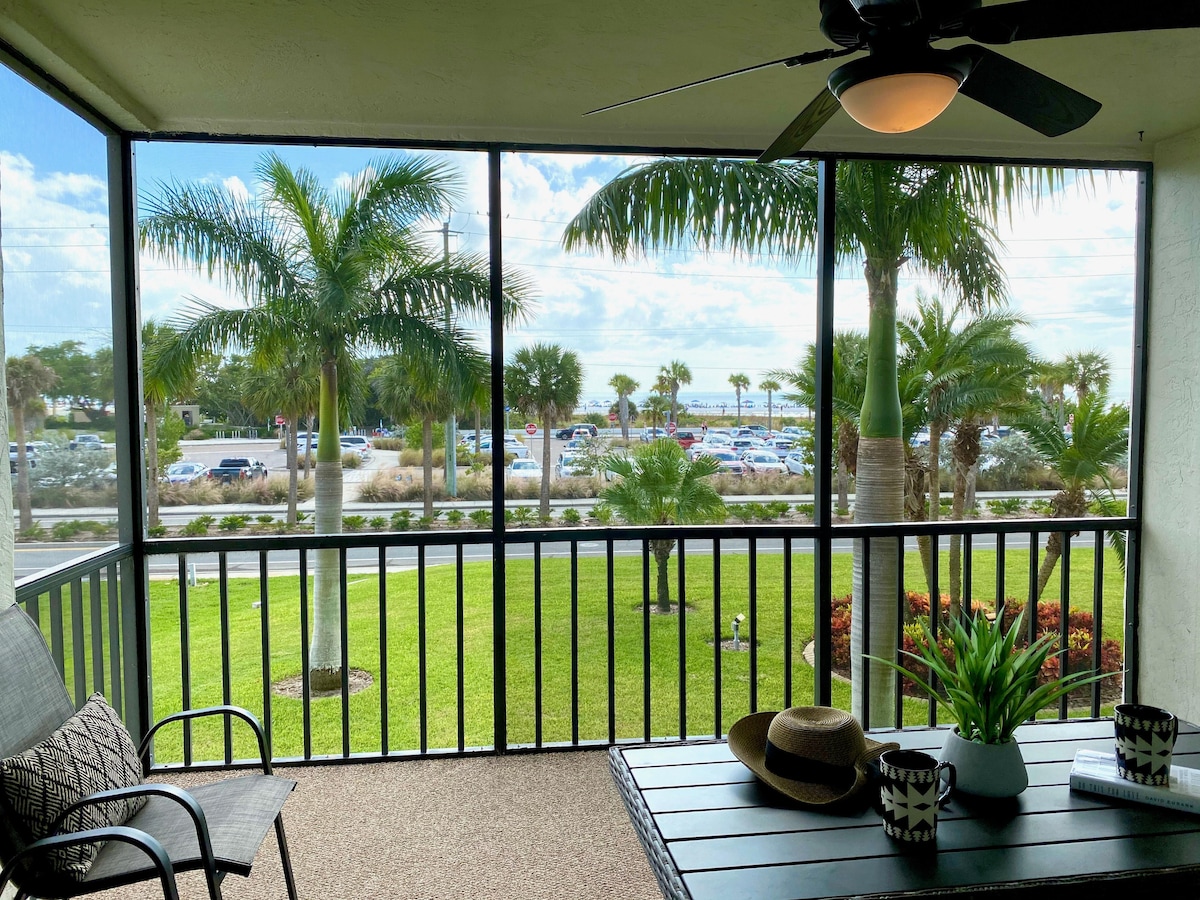 The screened lanai offers views of palm trees and well-maintained landscaping. A small table is positioned between two chairs, and a decorative hat rests on the table. A ceiling fan hangs above, providing a cooling breeze.
