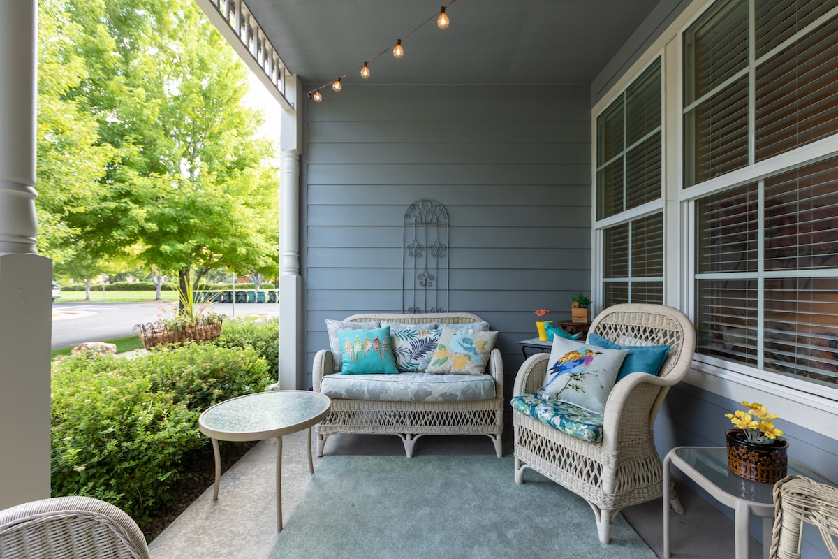 A cozy front porch is furnished with two wicker couches adorned with colorful cushions and a glass-topped table. Gentle lighting is provided by string lights overhead. Lush greenery surrounds the area, enhancing the welcoming atmosphere.