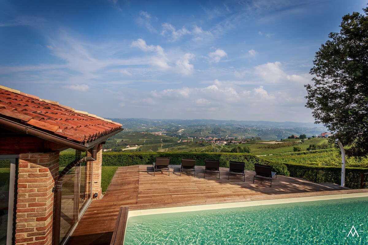The image captures the elegant swimming pool area surrounded by lush vineyards, showcasing a wooden sundeck with several sunbeds arranged for relaxation. Clear skies can be seen above, enhancing the expansive view of the rolling hills and vineyards in the distance.